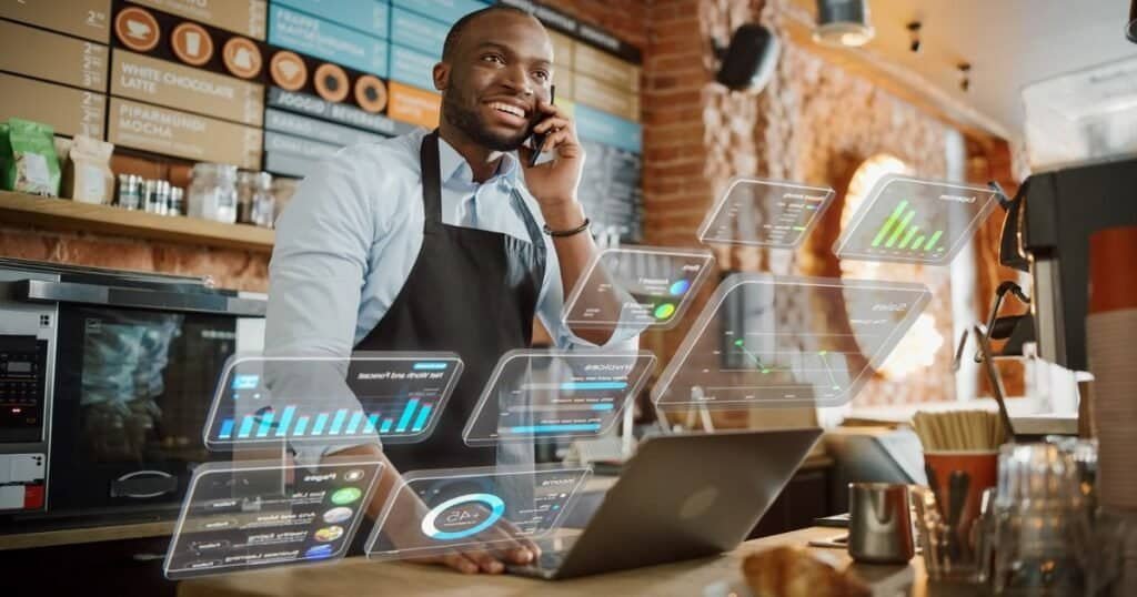 African American Coffee Shop Owner is Working on Computer and Talking on a Phone in a Cute Cafe. Augmented Reality Icons Popping Out of Restaurant Manager's Laptop: Small Black Business